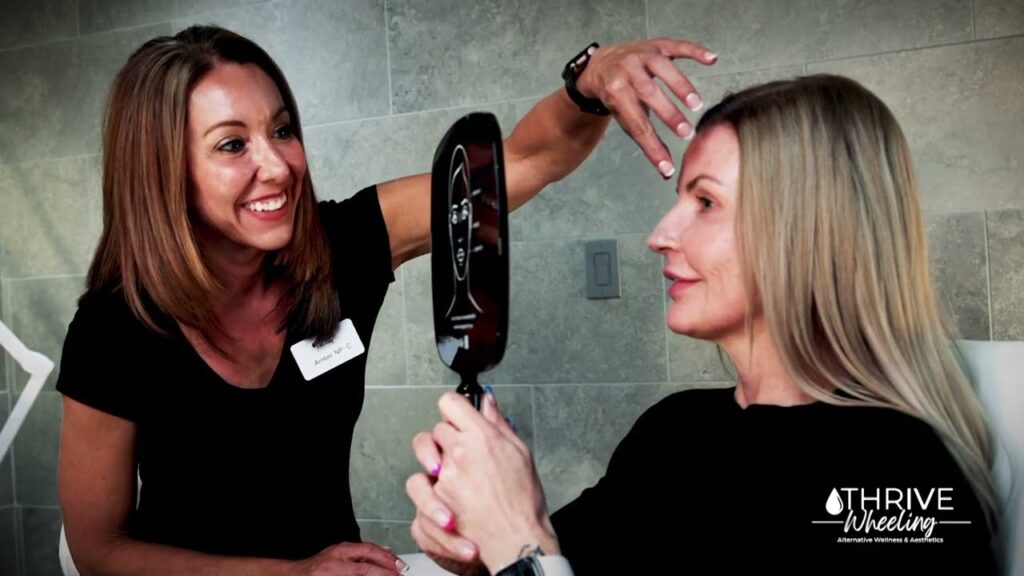 A provider at The Skin Clinic, smiling, works with a client to perform a facial assessment before an injection of Botox near St. Clairsville. The client, smiling as well, holds a hand mirror as the provider shows her different placement options on the forehead for Botox.