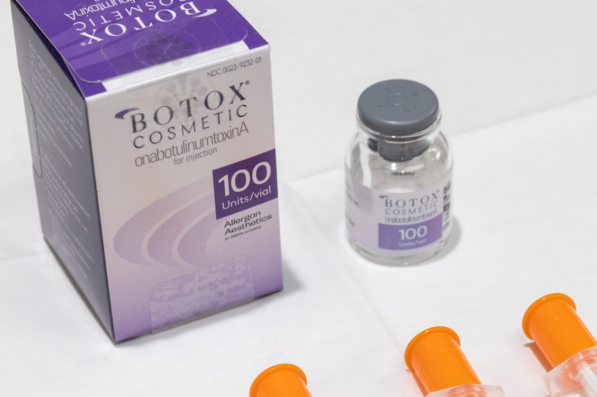 A vial of Botox near St. Clairsville sits on a white table alongside the box that the wrinkle relaxer comes in. At the bottom of the picture, the bright orange plunger covers of syringes are visible.